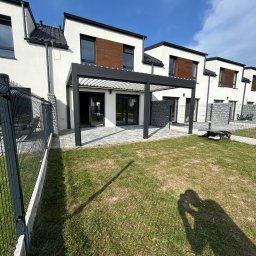 Elkano - Nowoczesna, czarna pergola metalowa przylegająca do domu z wyjściem na taras wyłożony szarą kostką. Widoczny fragment ogrodzenia i trawnik z cieniem osoby robiącej zdjęcie.