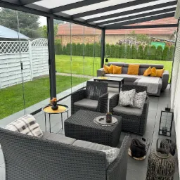 Zadaszony taras z meblami ogrodowymi: szary rattanowy zestaw wypoczynkowy, sofa z żółtymi poduszkami, lampiony. Widok na ogród z tujami i białe ogrodzenie.