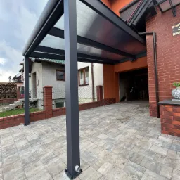 Nowoczesna, grafitowa pergola metalowa z przezroczystym zadaszeniem, przylegająca do budynku z cegły, z widoczną kostką brukową na podjeździe.