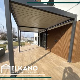 Elkano - Nowoczesna pergola z szarymi słupami i drewnianą ścianą, połączona z tarasem z płytek imitujących drewno. Widok na ogród w tle.