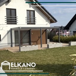 Elkano - Nowoczesna pergola z szarym stelażem i drewnianym panelem, zintegrowana z domem. Betonowe donice z roślinami na tarasie i trawnik przed domem.