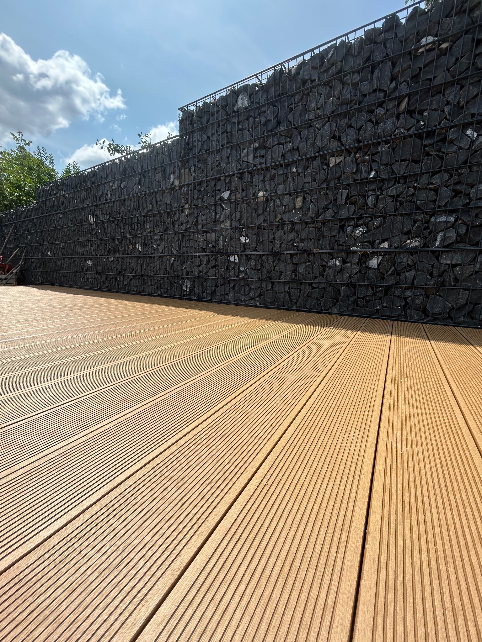 Nowoczesny taras kompozytowy z desek w odcieniach brązu, przylegający do gabionowej ściany z czarnych kamieni. Słoneczny dzień, błękitne niebo z chmurami.
