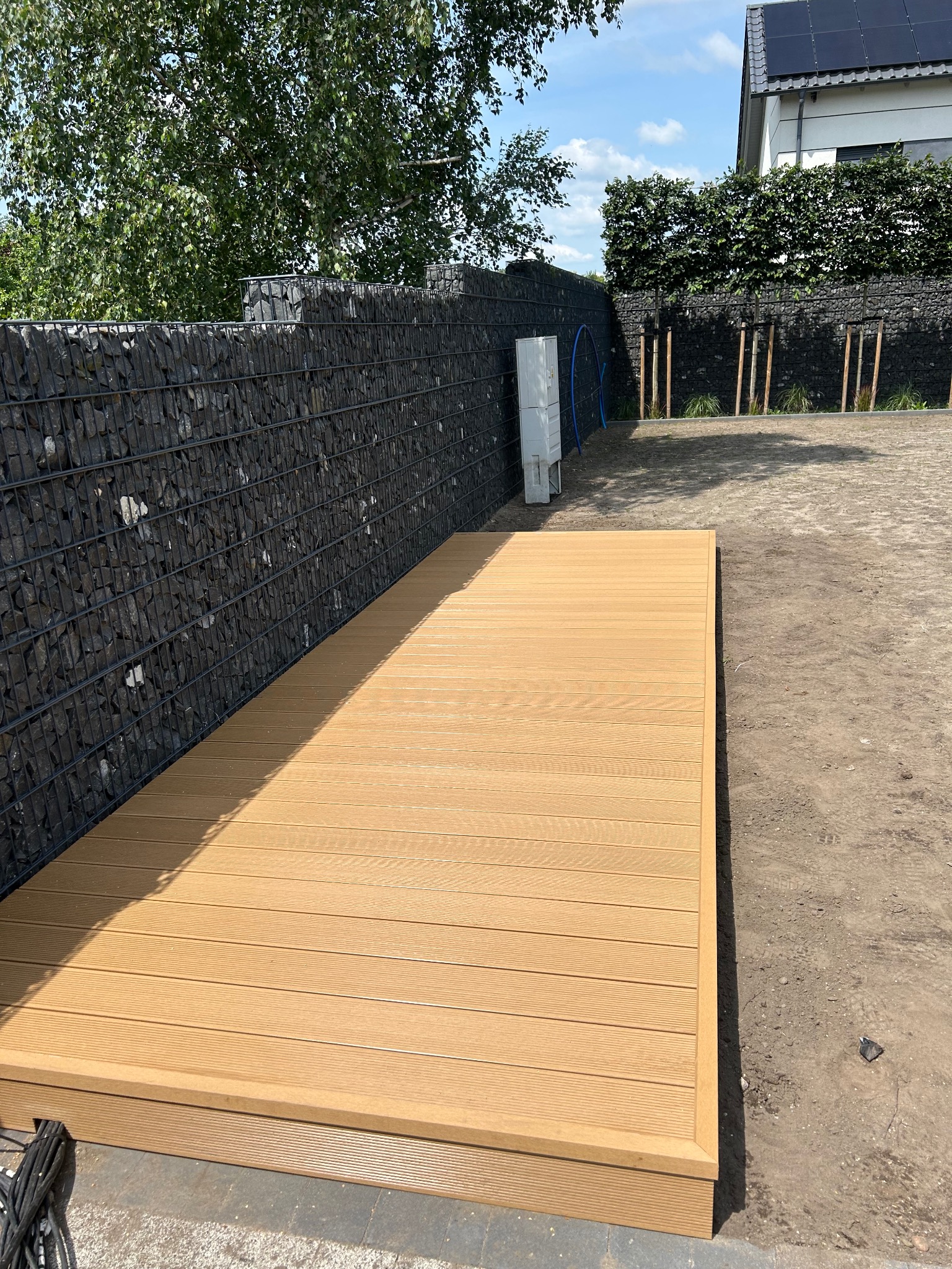 Nowy taras kompozytowy w odcieniach brązu, przylegający do gabionowego ogrodzenia. Widoczne przygotowane podłoże i elementy instalacji. Słoneczny dzień.