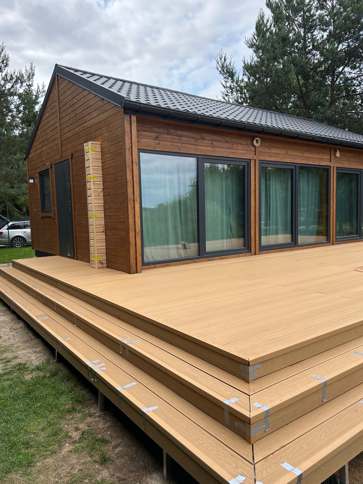 Nowoczesny dom z drewnianą elewacją i tarasem kompozytowym z widocznymi schodami, otoczony zielenią. Elementy wykończeniowe zabezpieczone taśmą.