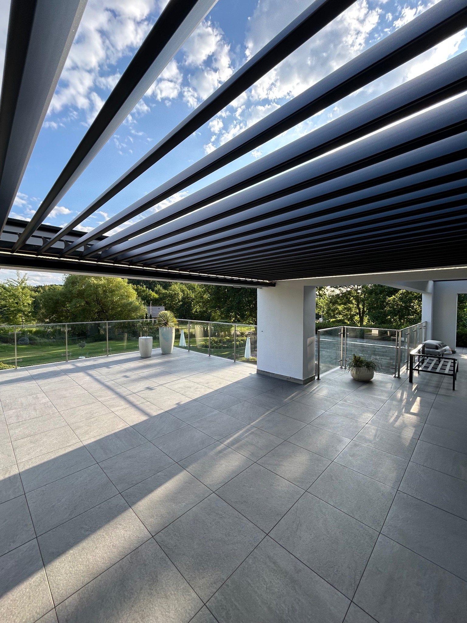 Nowoczesny taras z szarych płytek, zadaszony metalową pergolą z regulowanymi lamelami. Szklane balustrady, rośliny w donicach i meble ogrodowe dopełniają aranżację.