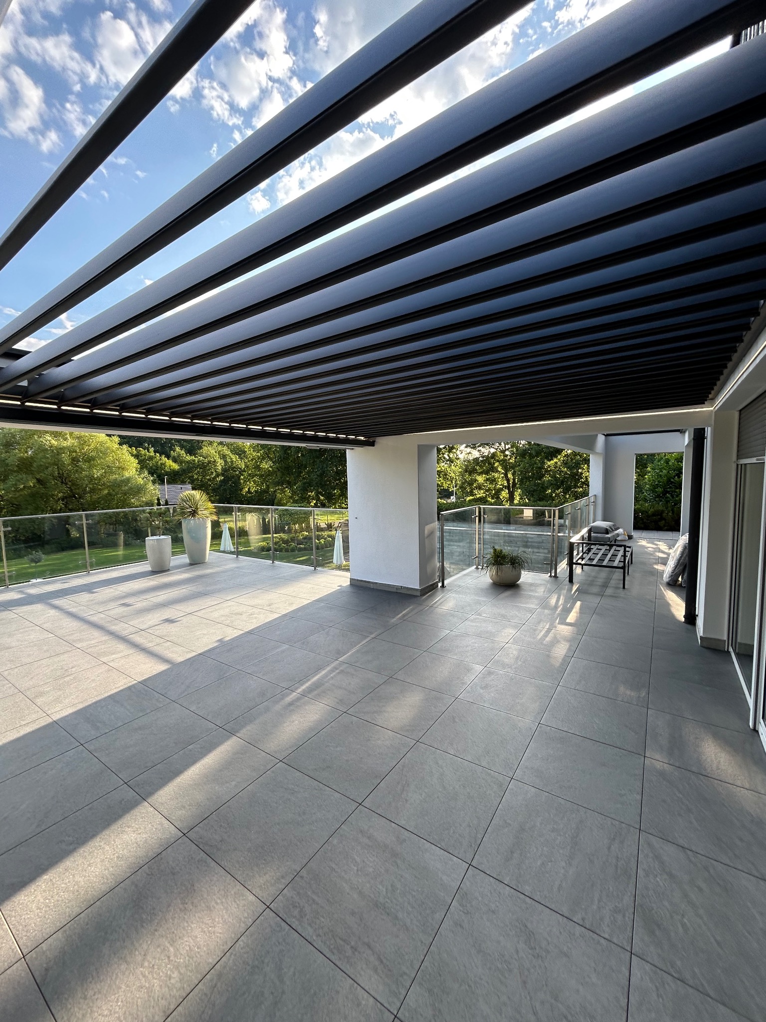 Nowoczesny taras z szarą, dużą płytką, zadaszony metalową pergolą z widokiem na zieleń. Szklane balustrady, minimalistyczne donice.