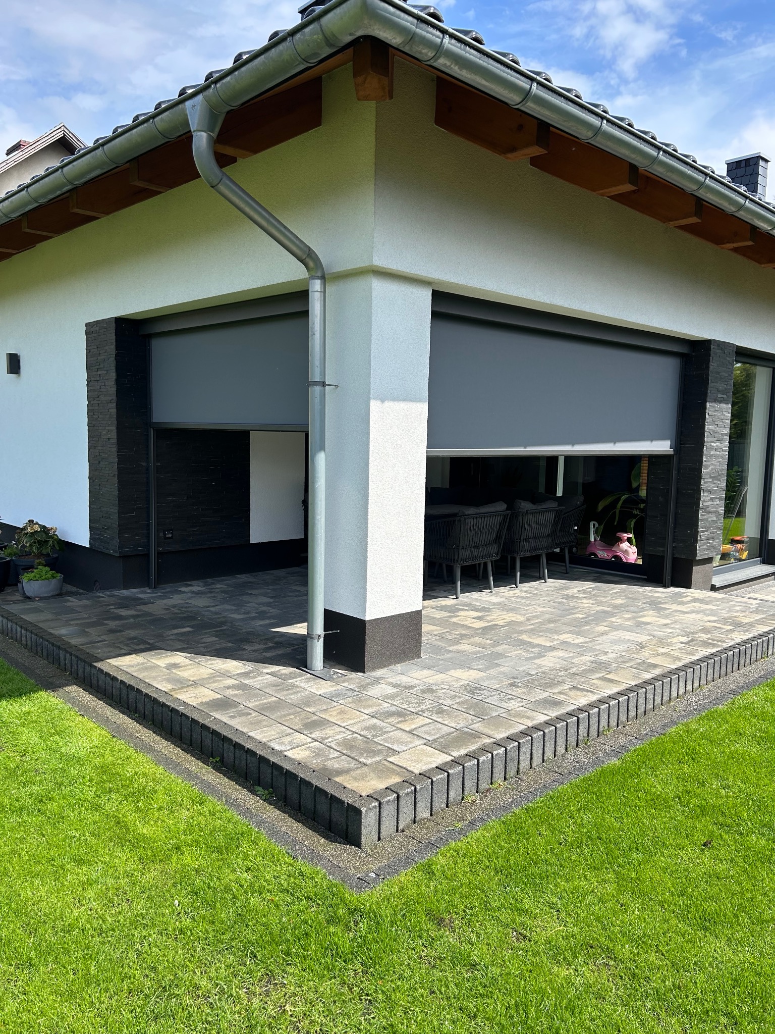 Nowoczesny taras z szarej kostki brukowej, obrzeża z ciemnych kostek, częściowo zasłonięty roletą zewnętrzną. Widok na meble ogrodowe i fragment trawnika.