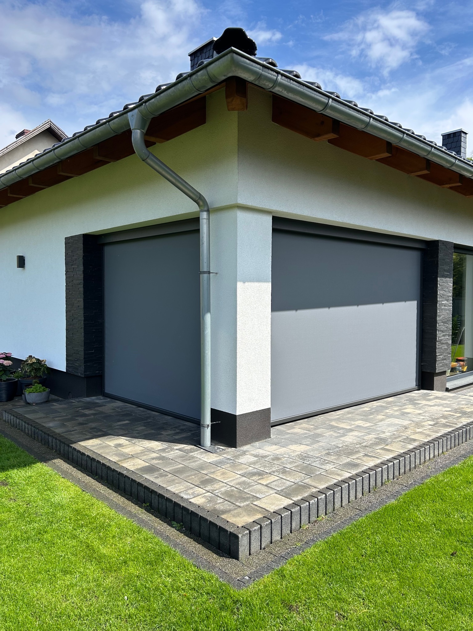 Szare rolety zewnętrzne na elewacji budynku z kostką brukową i zielonym trawnikiem w słoneczny dzień. Widoczny fragment dachu z rynną.