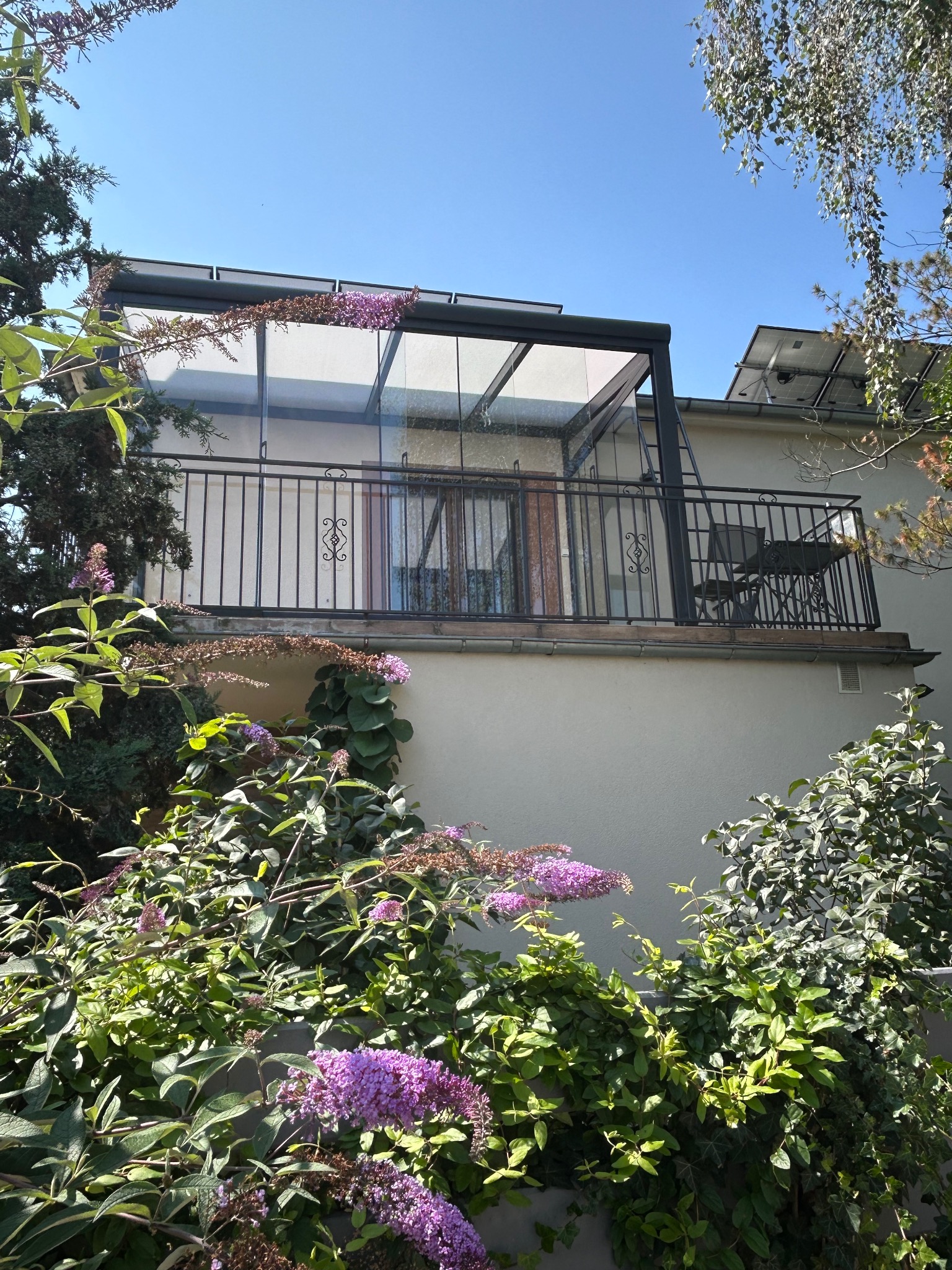 Zadaszony balkon z metalową balustradą, widoczny z dołu przez kwitnące krzewy. Na dachu budynku panele słoneczne. Balkon wyposażony w stolik i krzesła.