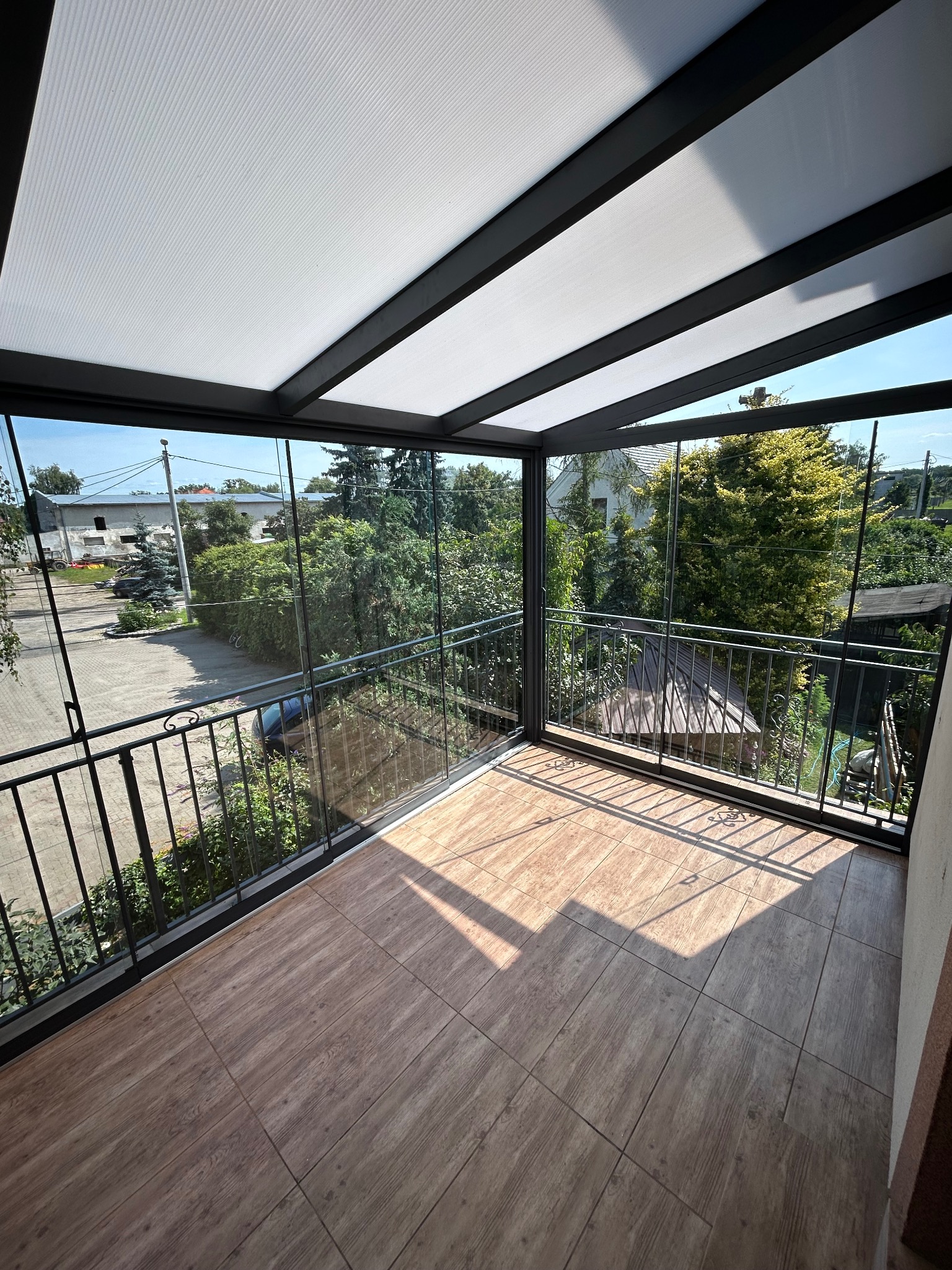 Zadaszony balkon z metalową konstrukcją i szklanymi ścianami, wykończony płytkami imitującymi drewno. Widok na zieleń i zabudowania z zewnątrz.