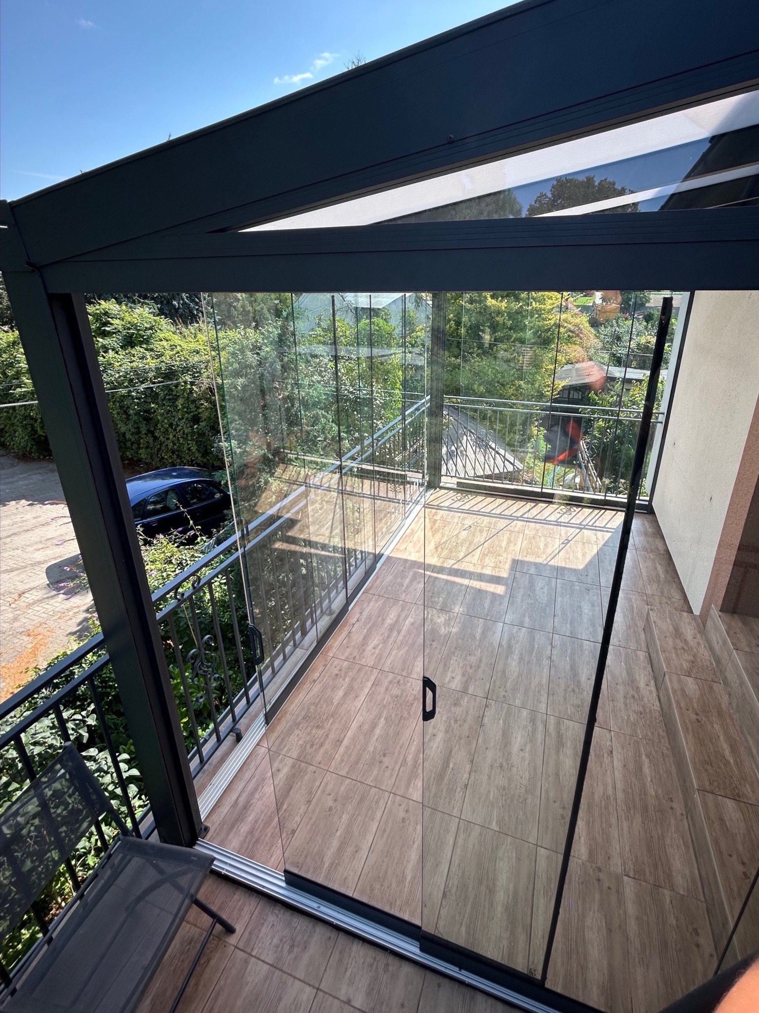 Zadaszony balkon z metalową ramą i szklanymi ścianami przesuwnymi. Podłoga wyłożona płytkami imitującymi drewno. Widok na zieleń i zabudowę.