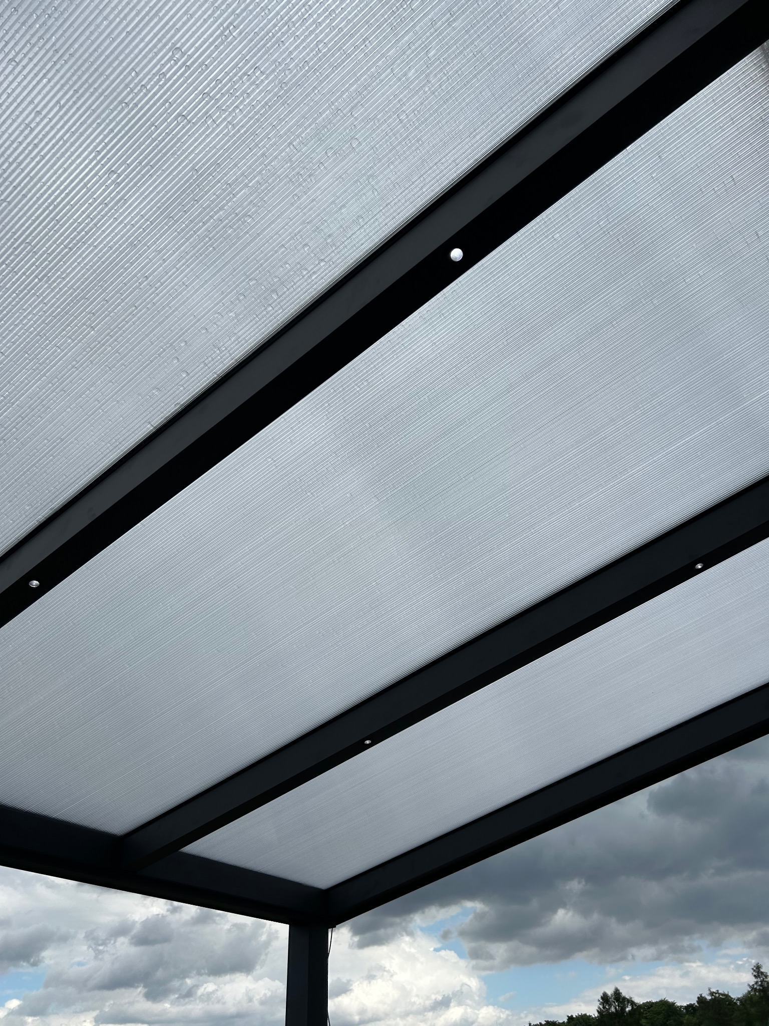 Nowoczesna pergola z czarną konstrukcją i przezroczystym zadaszeniem z widocznymi kroplami deszczu, widziana od dołu na tle pochmurnego nieba.