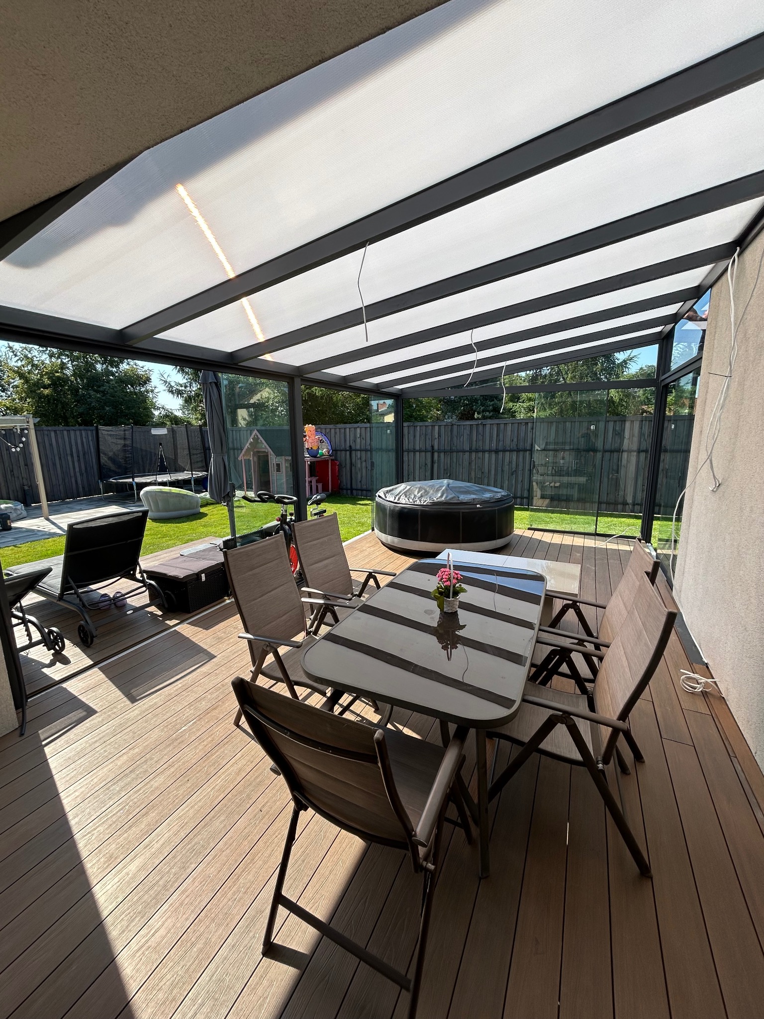 Zadaszony taras z kompozytową deską podłogową, stolikiem i krzesłami. W tle widoczny ogród z jacuzzi i zabawkami dla dzieci. Nowoczesna konstrukcja z białym zadaszeniem.