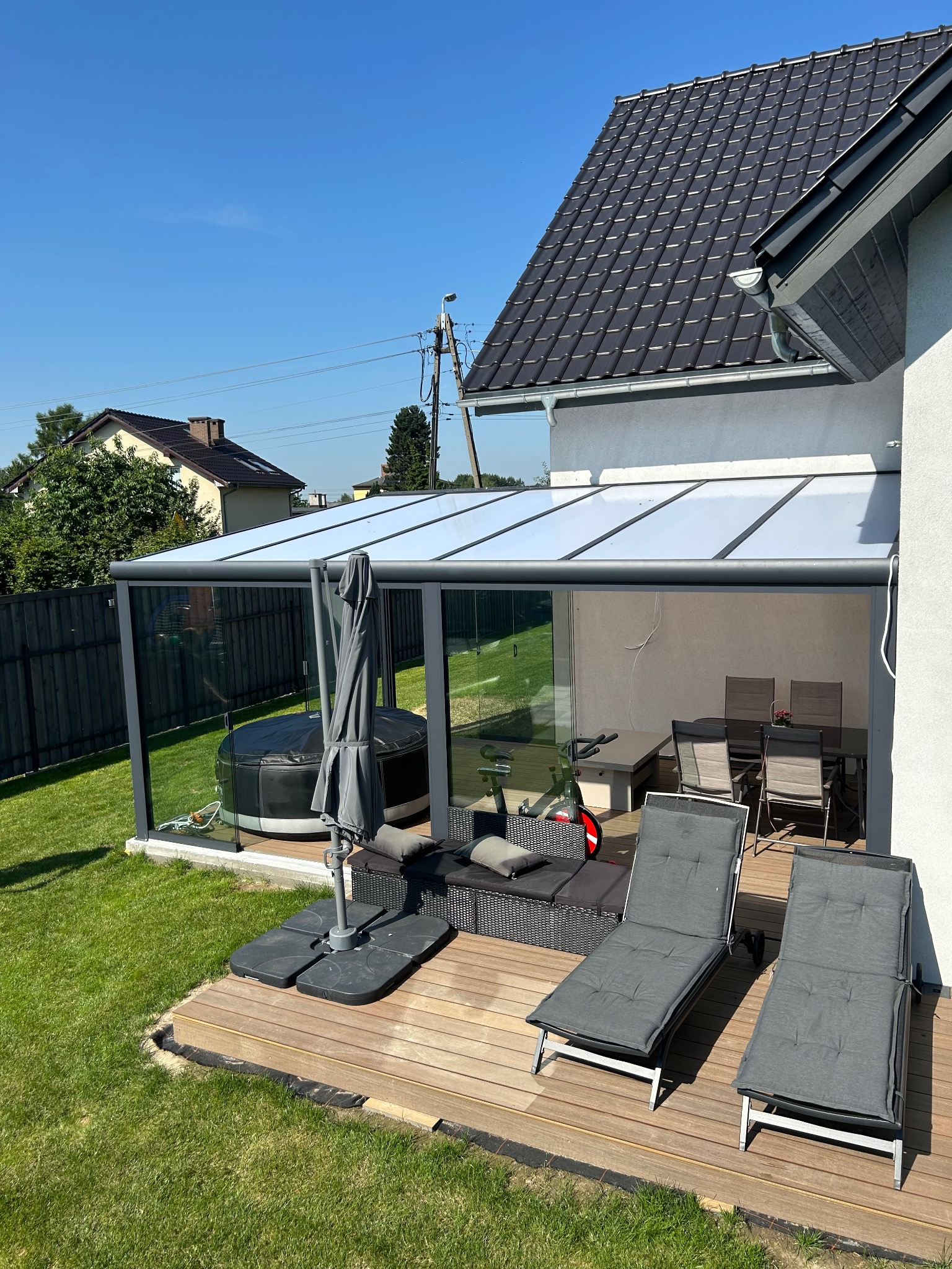 Zadaszony taras z drewnianą podłogą, wyposażony w jacuzzi, leżaki i stół z krzesłami. Widok na ogród i dom w tle, słoneczny dzień.