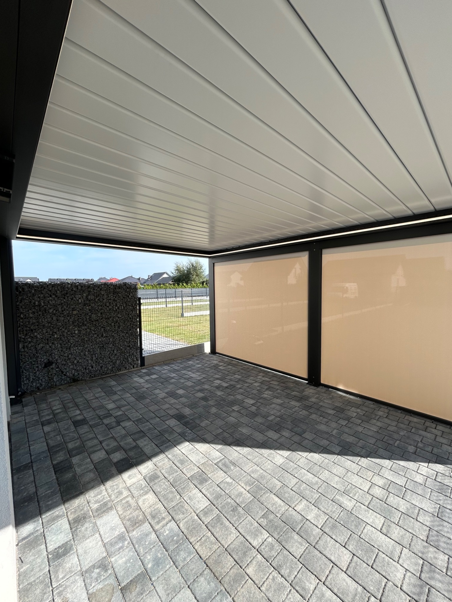 Zadaszony taras z szarą kostką brukową i beżowymi roletami bocznymi. Widoczny fragment gabionu z kamienia. Oświetlenie LED pod dachem.