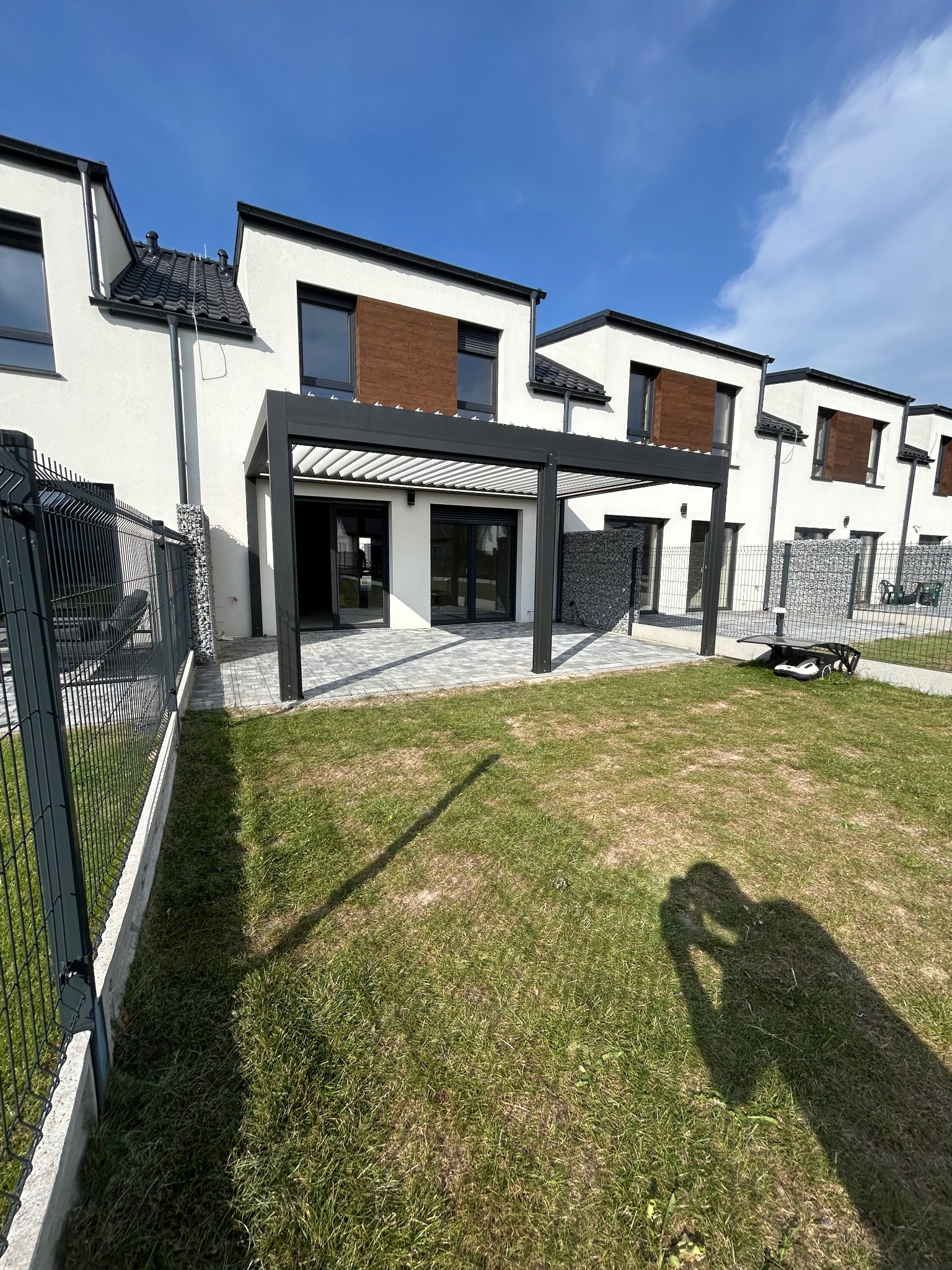 Nowoczesna, czarna pergola metalowa przylegająca do domu z wyjściem na taras wyłożony szarą kostką. Widoczny fragment ogrodzenia i trawnik z cieniem osoby robiącej zdjęcie.