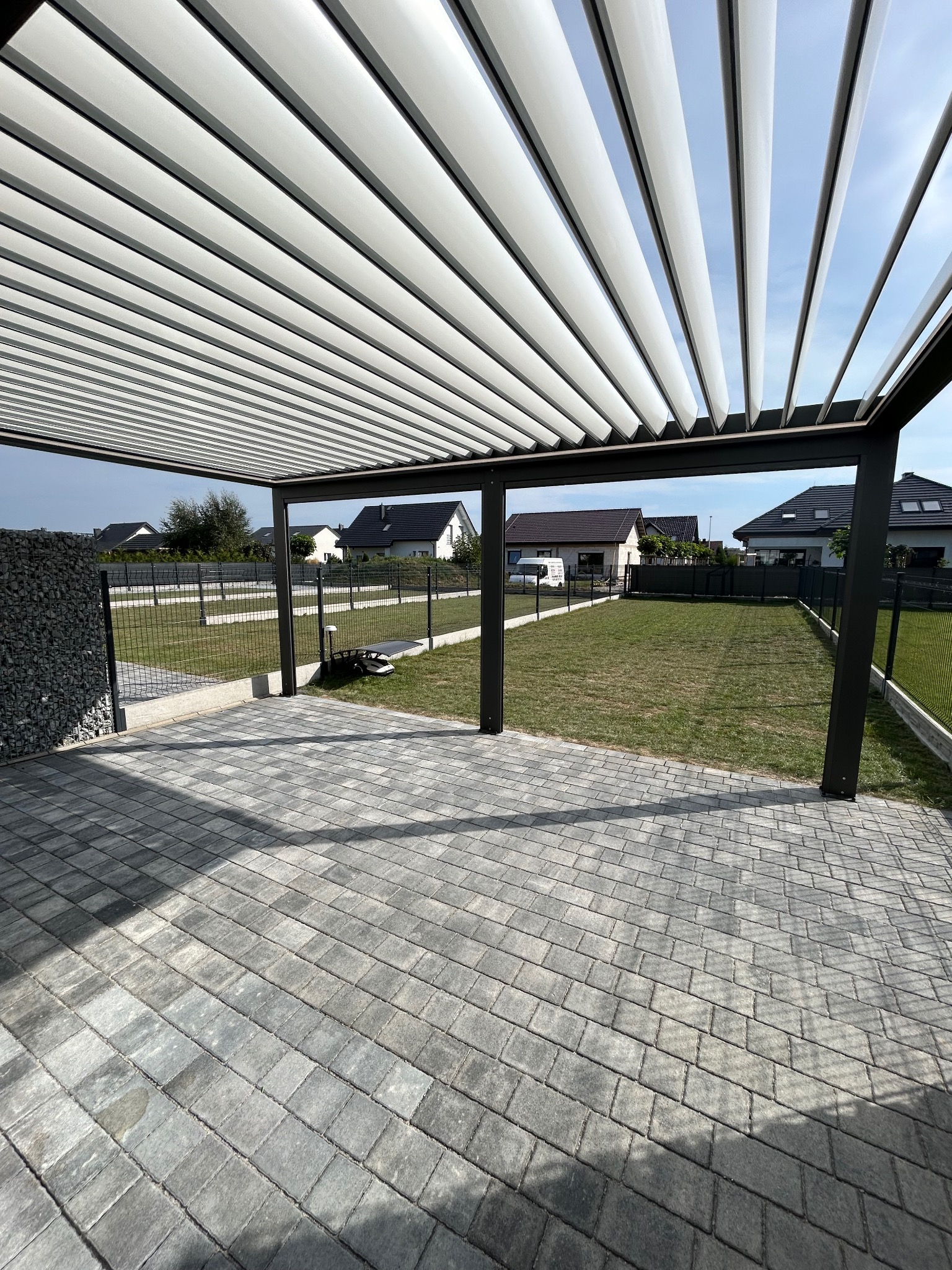 Nowoczesna pergola metalowa z regulowanymi lamelami, zacieniająca taras z szarej kostki brukowej. W tle widoczne domy jednorodzinne i ogrodzenie.