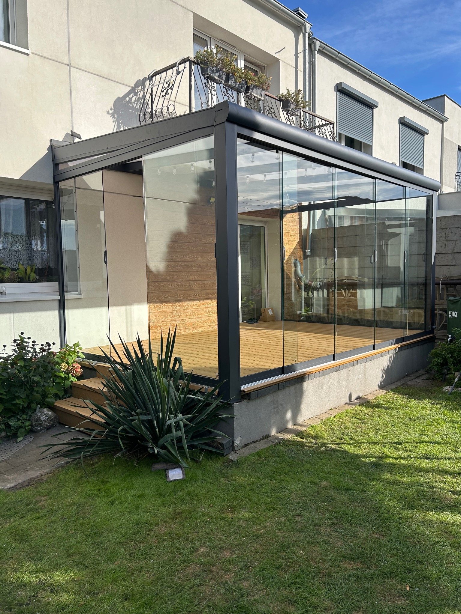 Szklana zabudowa tarasu z drewnianą podłogą, widok z zewnątrz, zieleń w tle, budynek z oknami i balkonem. Nowoczesna konstrukcja, jasne drewno, schludny ogród.