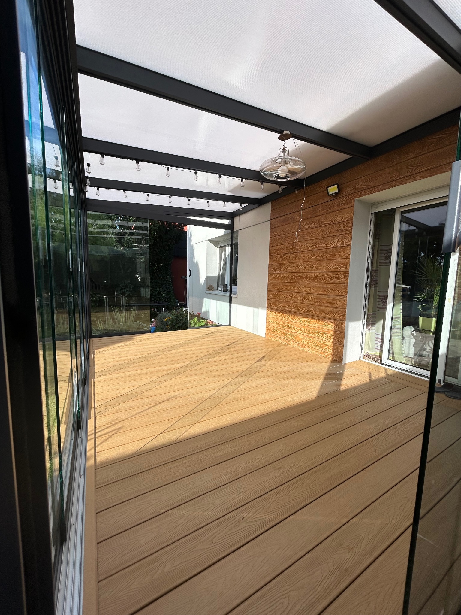 Zadaszony taras z kompozytową podłogą, oświetleniem i szklanymi panelami bocznymi przylegający do budynku z drewnianą elewacją i dużymi oknami.