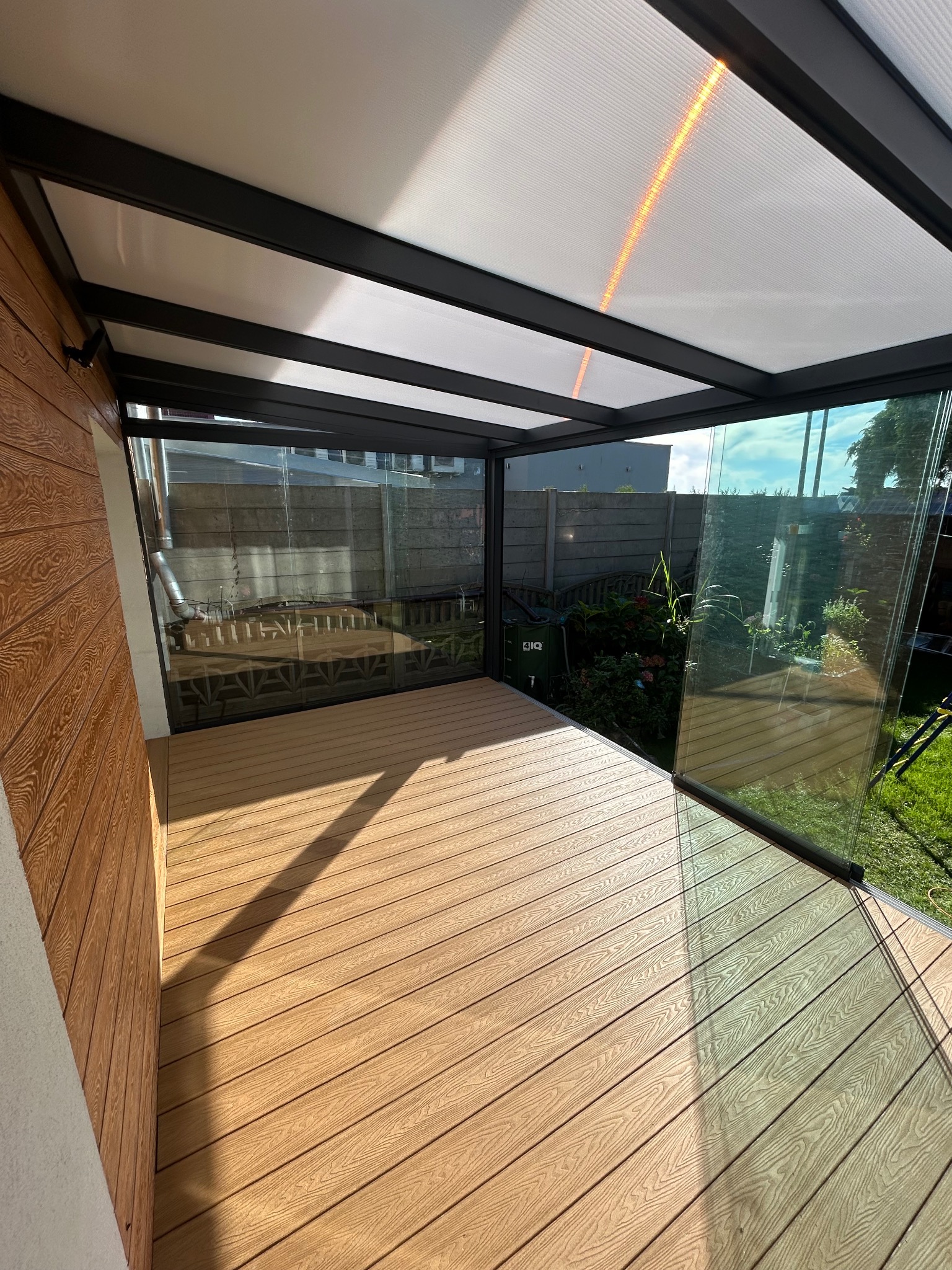 Zadaszony taras z kompozytową podłogą i szklanymi ścianami, widok na ogród. Nowoczesna konstrukcja z czarną ramą i drewnianą elewacją.