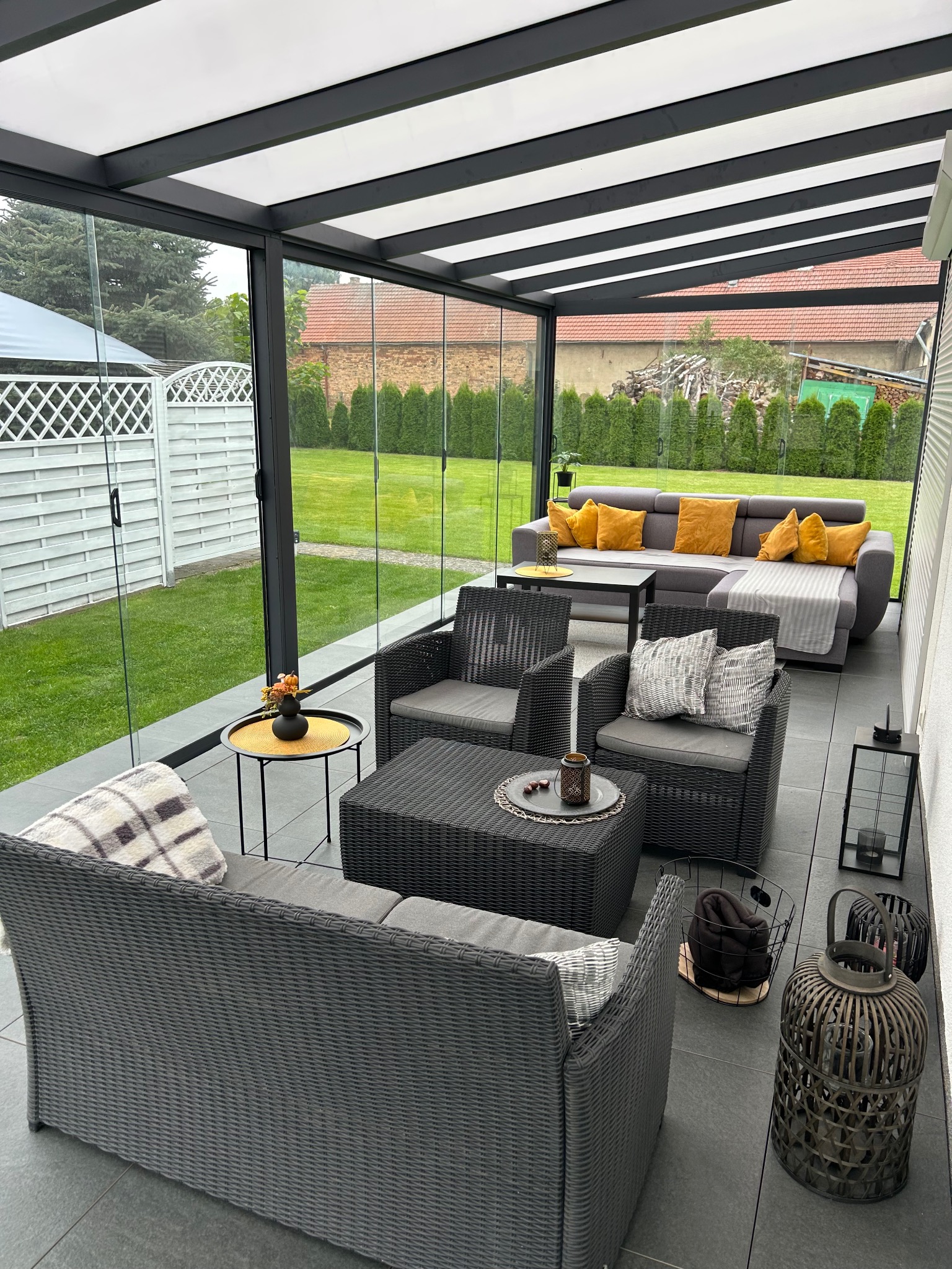 Zadaszony taras z meblami ogrodowymi: szary rattanowy zestaw wypoczynkowy, sofa z żółtymi poduszkami, lampiony. Widok na ogród z tujami i białe ogrodzenie.