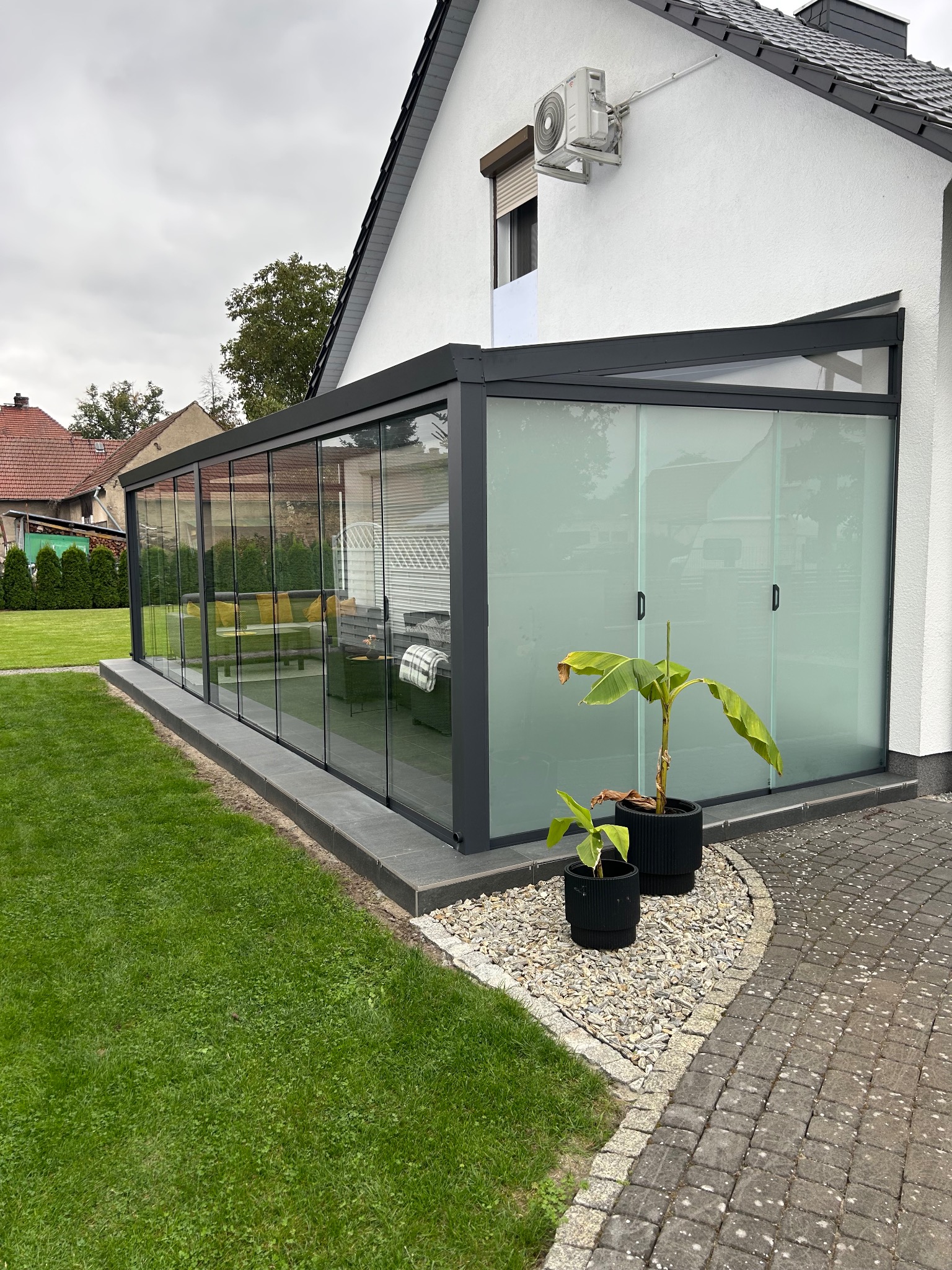 Nowoczesna pergola z przesuwnymi szklanymi ścianami, z czarną ramą, przylegająca do białego domu. Dwie donice z roślinami stoją na żwirowym podłożu.