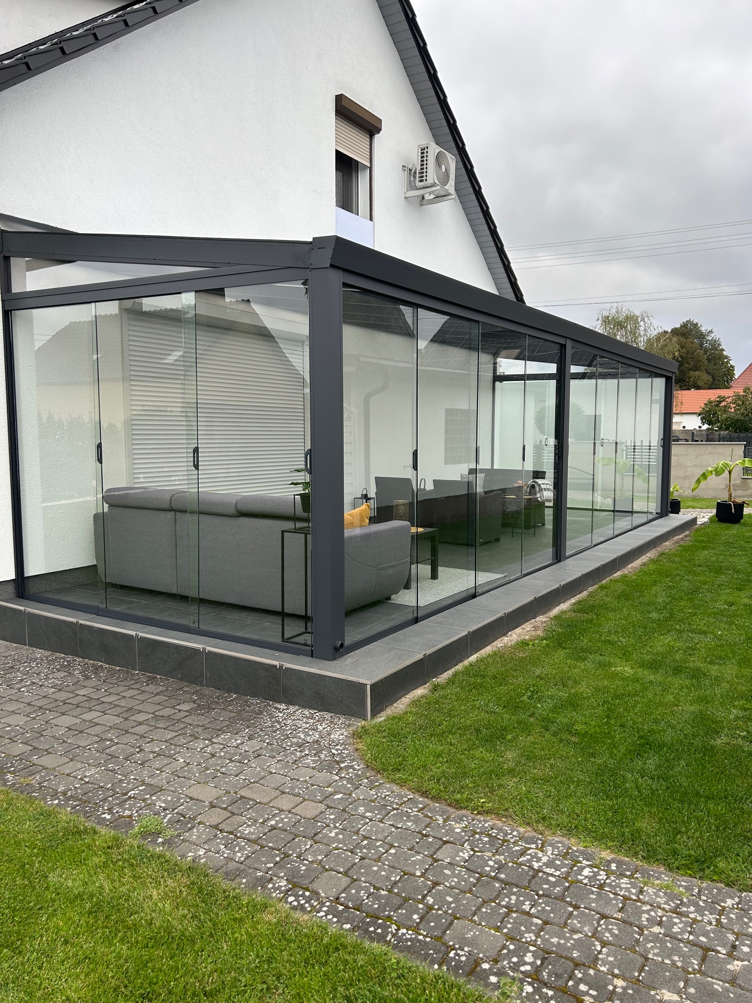 Szklana pergola z przesuwnymi drzwiami, szara konstrukcja, widok z zewnątrz na salonik z sofami. Płytki na tarasie i trawnik. Nowoczesny design.