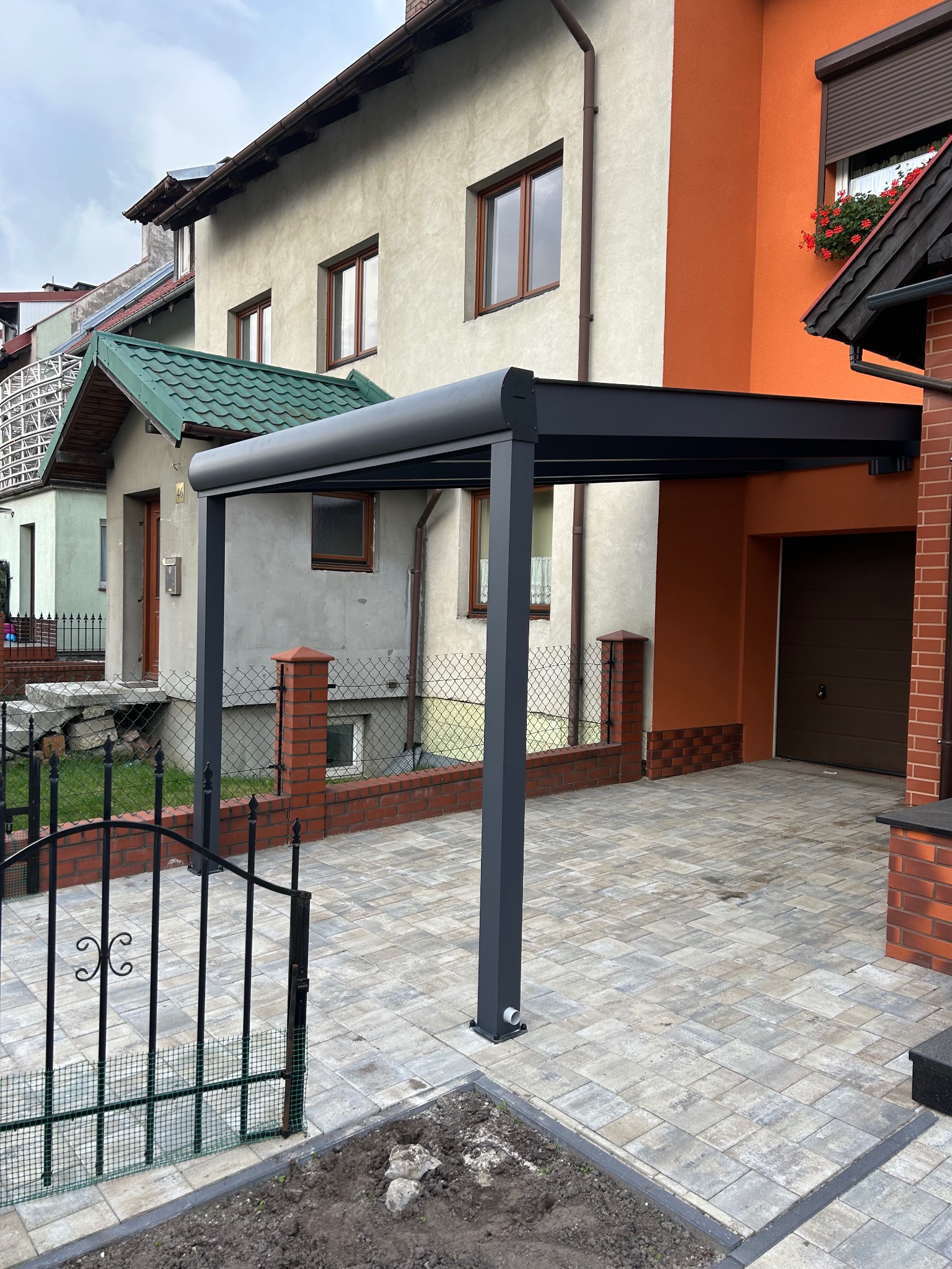 Nowoczesna, grafitowa pergola metalowa przy domu z brukowanym podjazdem. Widoczne elementy ogrodzenia i sąsiednie budynki, pochmurne niebo w tle.