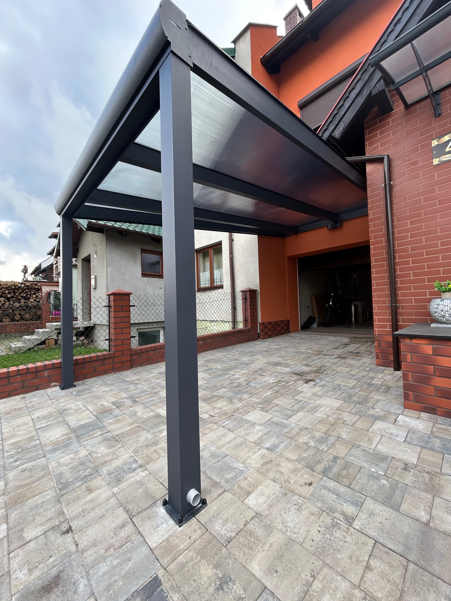 Nowoczesna, grafitowa pergola metalowa z przezroczystym zadaszeniem, przylegająca do budynku z cegły, z widoczną kostką brukową na podjeździe.