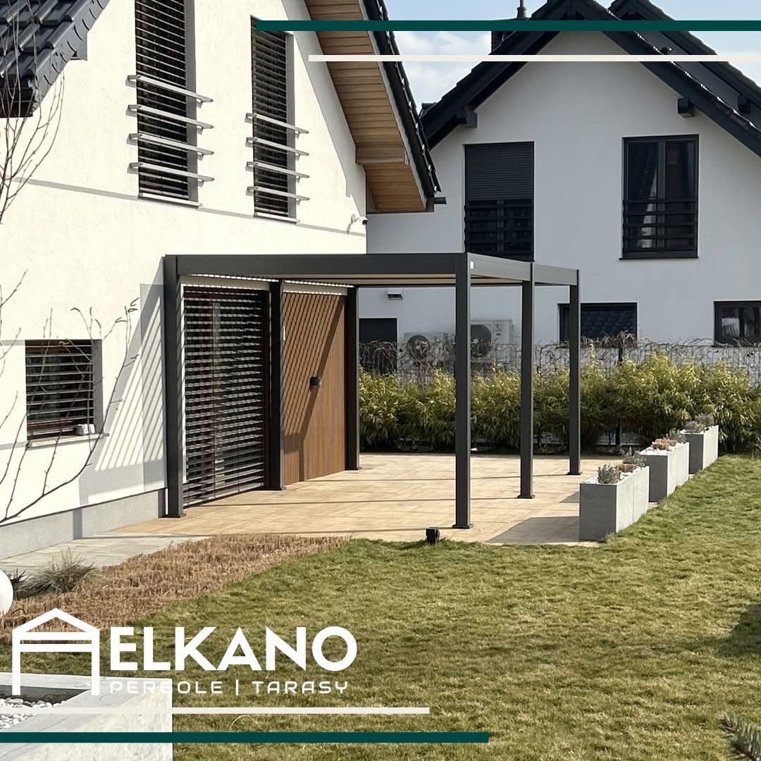 Nowoczesna pergola z szarym stelażem i drewnianymi panelami, przylegająca do białego domu z żaluzjami. Uwagę zwraca jasna, kamienna posadzka tarasu i minimalistyczne donice.