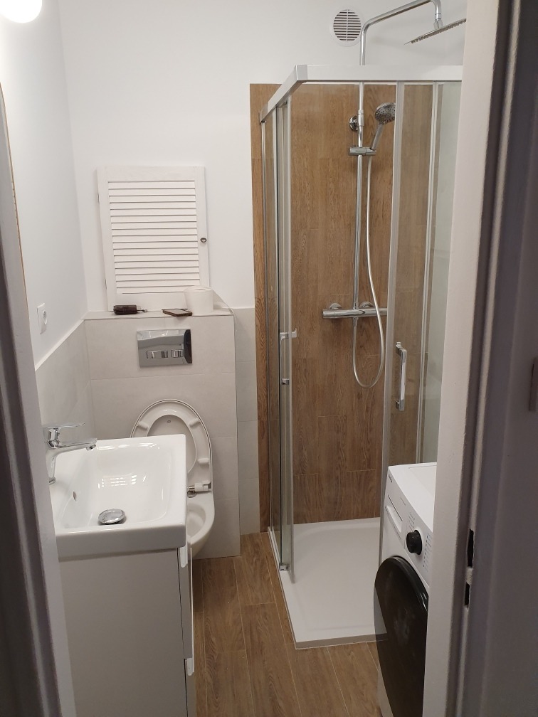 Nowoczesna, jasna łazienka po remoncie w Poznaniu: kabina prysznicowa z panelami imitującymi drewno, biała umywalka z szafką i toaleta z zabudową, pralka w rogu.