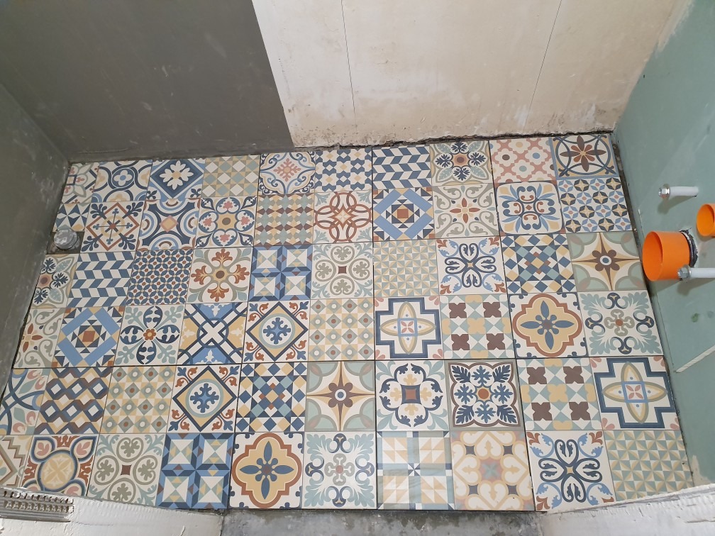 Ułożone, wzorzyste płytki podłogowe w łazience w trakcie remontu, widoczne fragmenty ścian w surowym stanie i pomarańczowe rury kanalizacyjne.