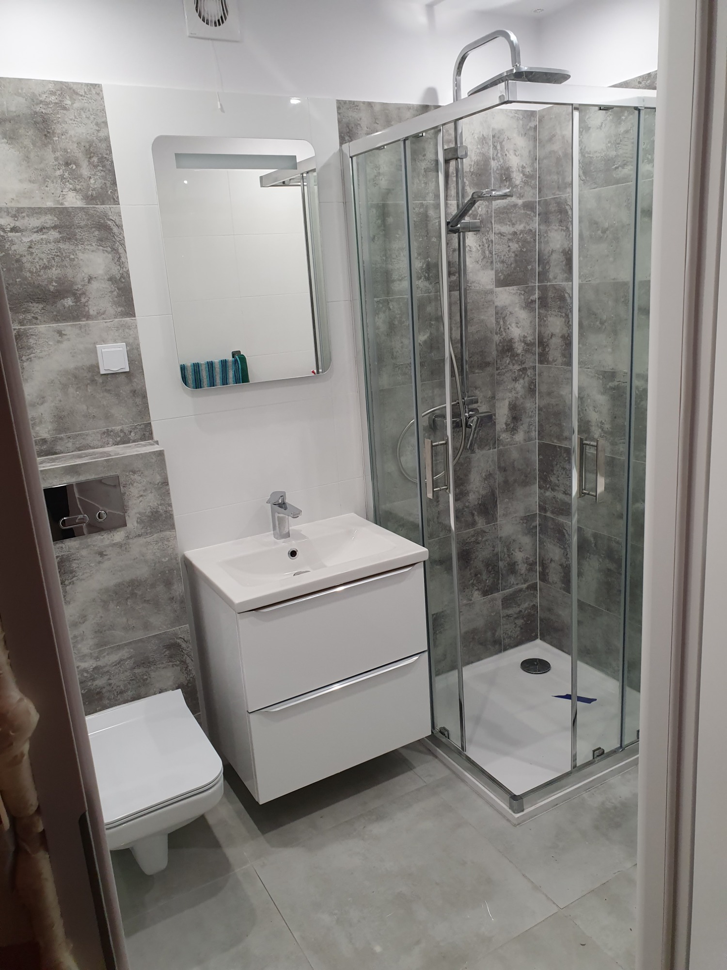 Nowoczesna łazienka z kabiną prysznicową, umywalką z szafką i podwieszaną toaletą, wykończona szarymi płytkami imitującymi kamień.