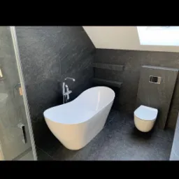 Nowoczesna łazienka w odcieniach szarości z wolnostojącą białą wanną, szklaną kabiną prysznicową i podwieszaną toaletą na tle ściany z ciemnego kamienia.