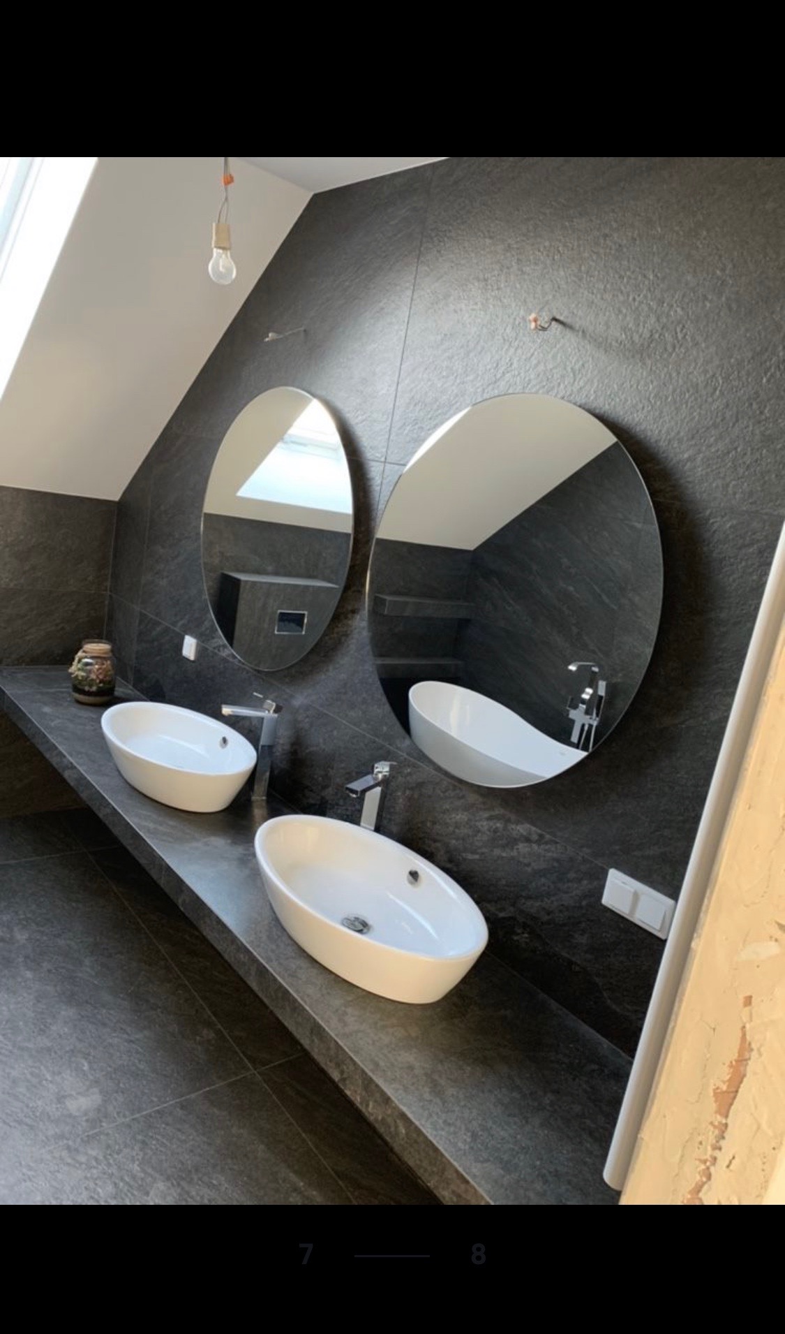 Nowoczesna łazienka na poddaszu z dwoma owalnymi umywalkami nablatowymi, dużymi okrągłymi lustrami i ciemnymi płytkami imitującymi kamień na ścianach i podłodze.