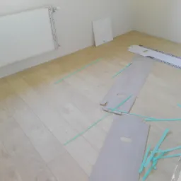 Jasne panele podłogowe ułożone w pomieszczeniu, widoczne fragmenty opakowań paneli Kronopol Platinum Flooring i zielone kliny dystansowe.