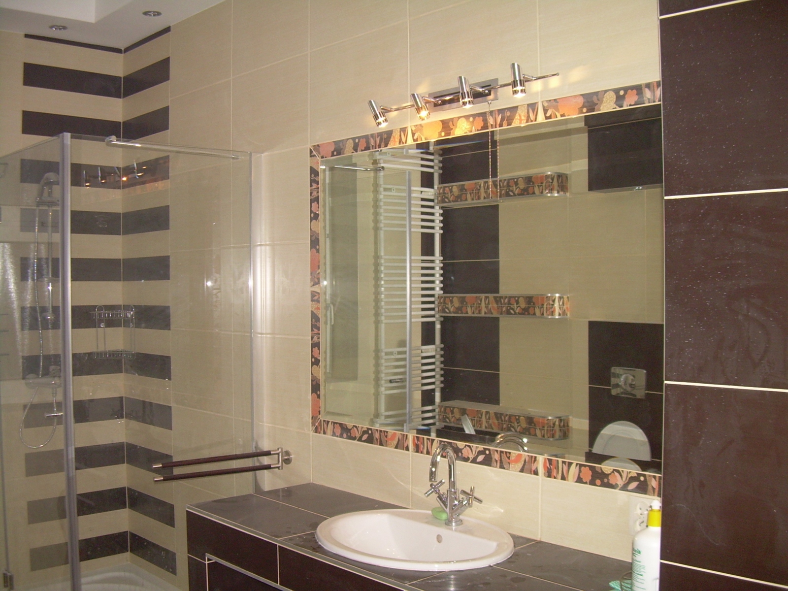 Wykończona łazienka z kabiną prysznicową, umywalką w blacie z ciemnego konglomeratu i dużym lustrem z ozdobną ramą, oświetlona halogenami.