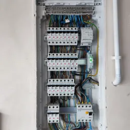 Otwarta rozdzielnica elektryczna wnękowa z zainstalowanymi bezpiecznikami EATON, modułem Zamel TRM-12 i zielonym wskaźnikiem LED LK-713, widoczne kable w różnych kolorach, ściana w stanie surowym.