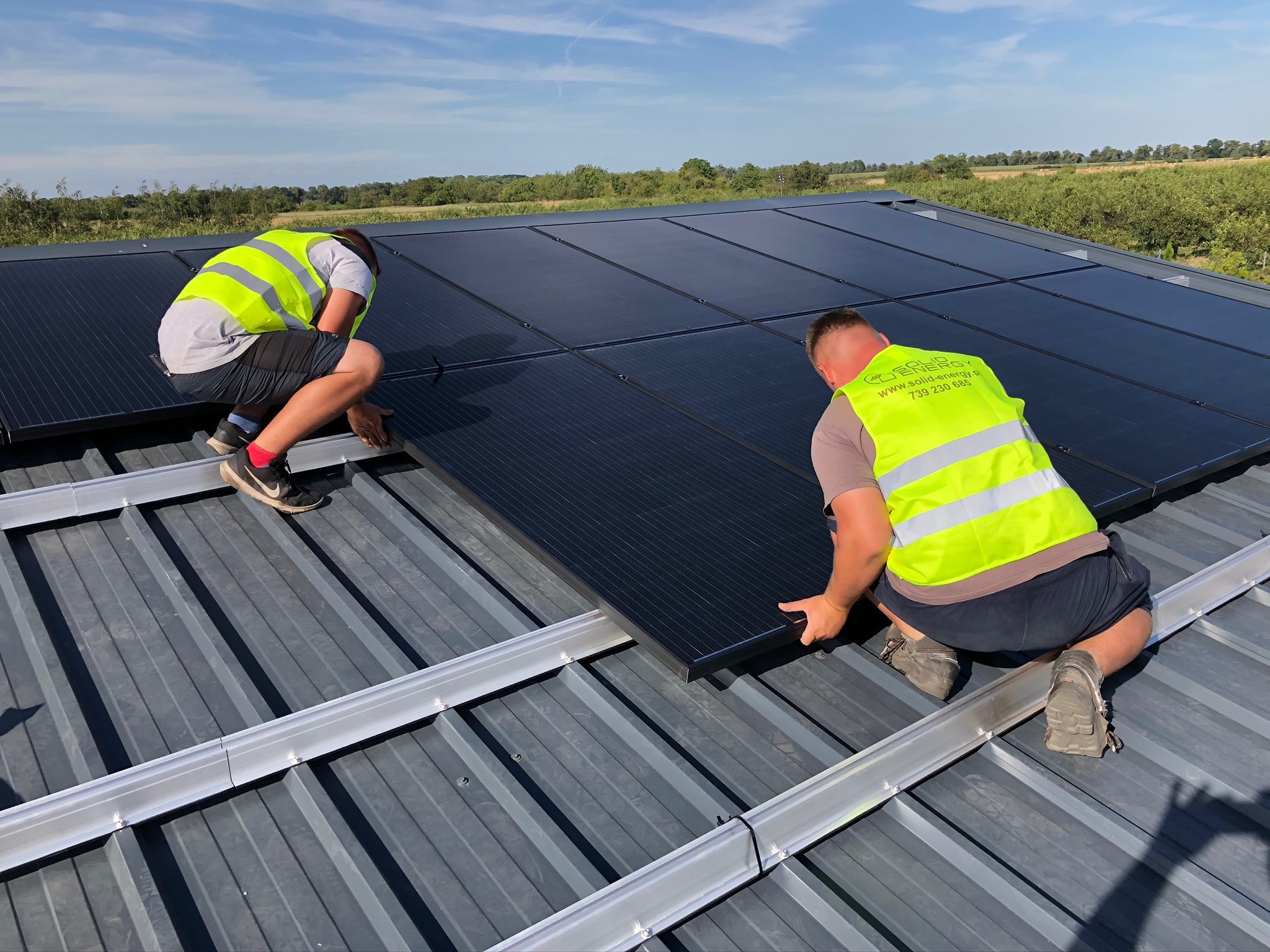 Dwóch instalatorów w jaskrawych kamizelkach montuje panele słoneczne na metalowym dachu, widok z góry na rozległy krajobraz.