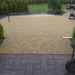 Żółta kostka brukowa ułożona na podjeździe, z widoczną zagęszczarką i elementami dekoracyjnymi z ciemnej kostki imitującej drewno, otoczona zielenią.