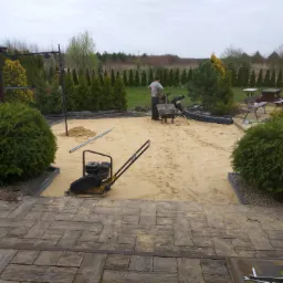 Przygotowanie podłoża pod taras kamienny: ubijarka spalinowa na piasku, widoczne obrzeża, narzędzia i pracownicy w tle, otoczenie z tujami i elementami architektury ogrodowej.