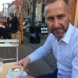 Mężczyzna w białej koszuli siedzi w kawiarni na zewnątrz, trzyma filiżankę cappuccino z wzorem serca na piance, w tle widoczny budynek z czerwonej cegły.