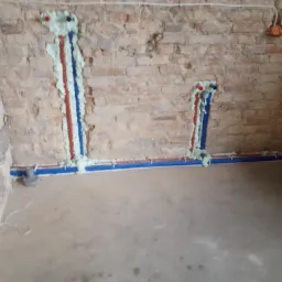 Miedziane i niebieskie rury wodne w ścianie z czerwonej cegły, zabezpieczone pianką montażową, widoczne na tle surowego betonu.