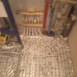 Rozdzielacz podłogowy z licznymi pętlami rur grzewczych ułożonych na folii izolacyjnej, widoczne elementy instalacji CO na ścianie.