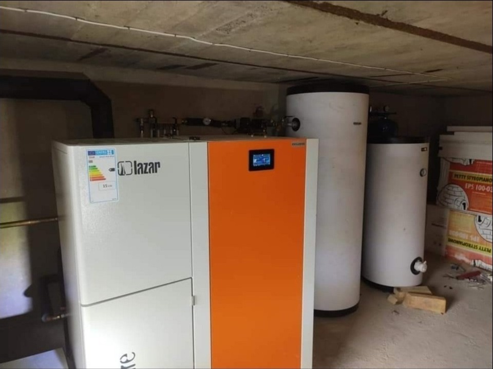 Kocioł na pellet Lazar Exklusiv z panelem sterowania i zbiornikami buforowymi w kotłowni, widoczny fragment instalacji grzewczej i opakowania styropianu.