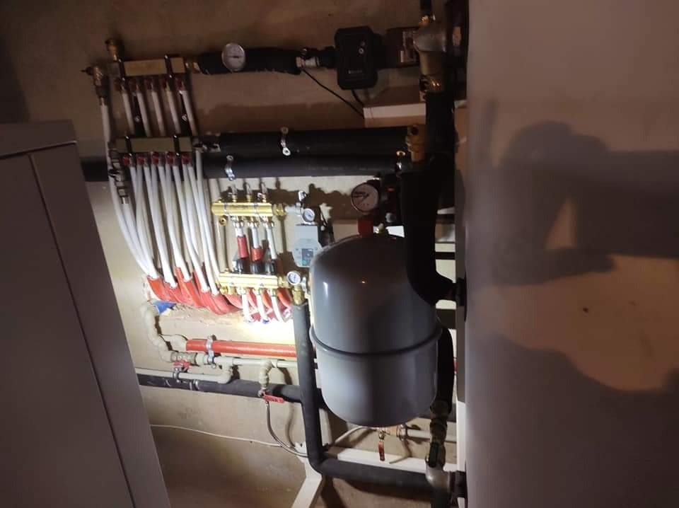 Rozdzielacz instalacji ogrzewania podłogowego z białymi rurami w ścianie, zbiornik wyrównawczy i liczne zawory oraz manometry w kotłowni.