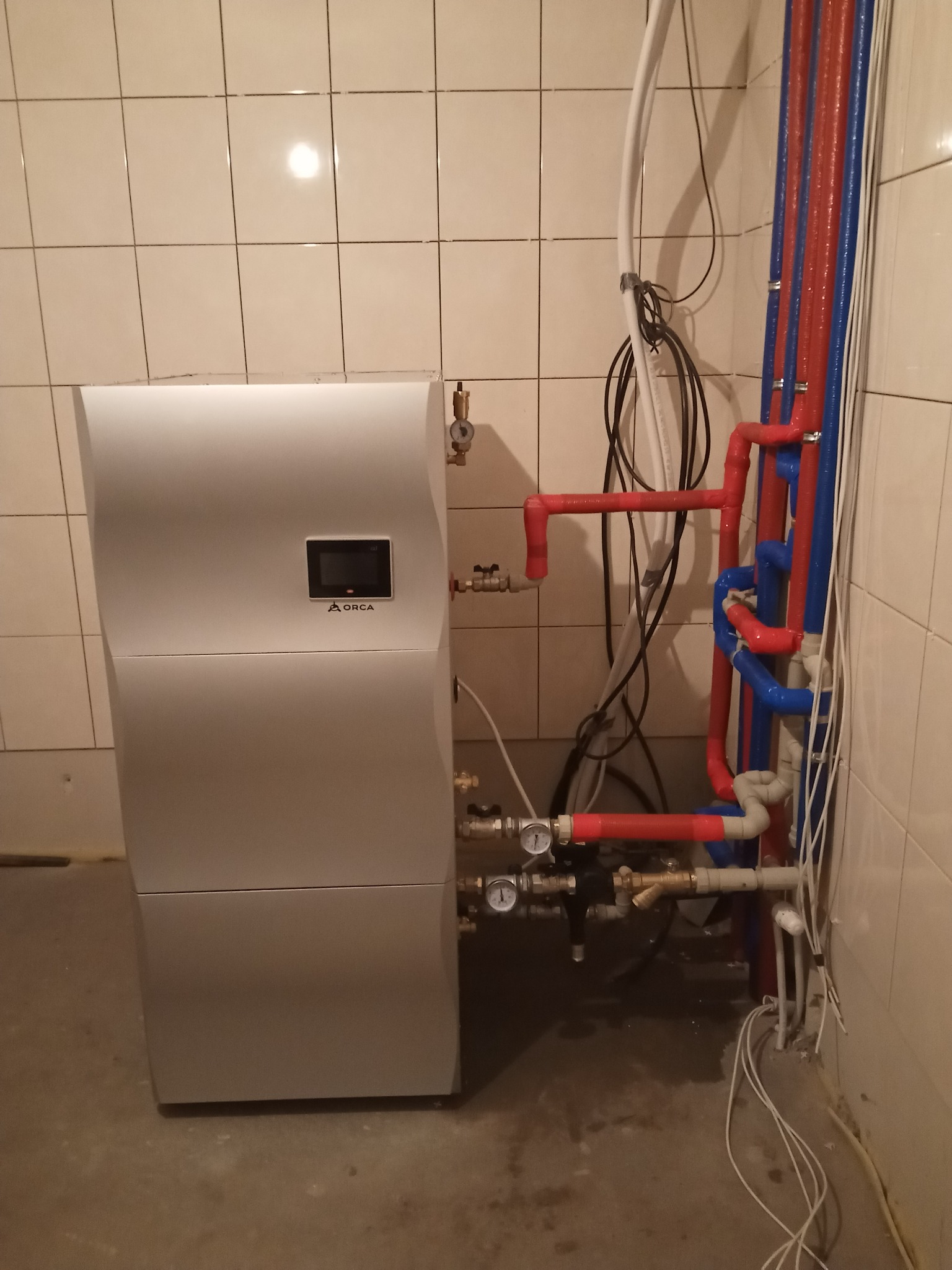 Nowoczesna pompa ciepła Mitsubishi Orca typu split, z widocznymi podłączeniami rur w kolorach czerwonym i niebieskim, zamontowana w pomieszczeniu z wykafelkowanymi ścianami.