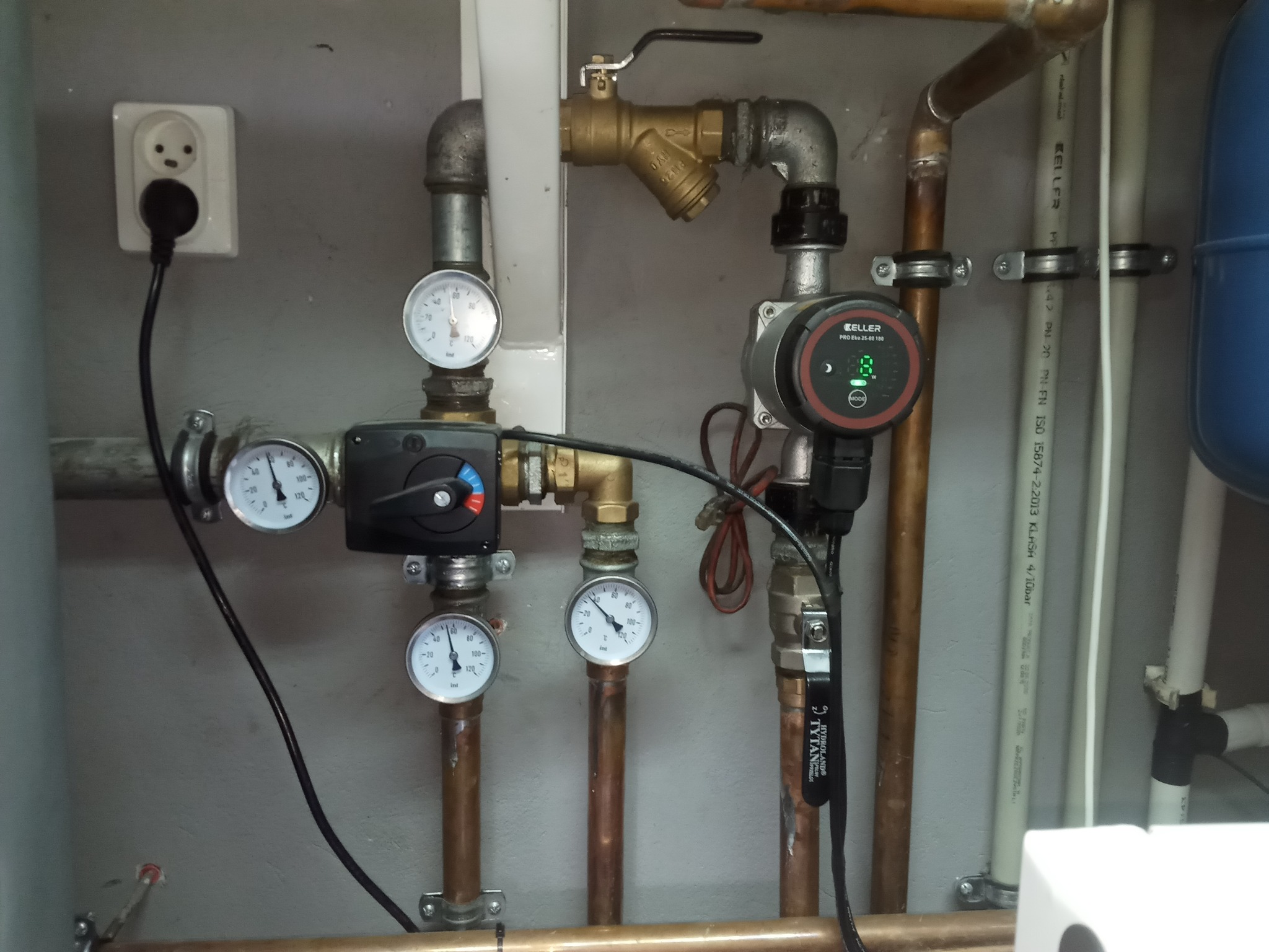 Fragment instalacji z miedzianymi rurami, manometrami, pompą obiegową Keller PRO Eko 25-60 180 oraz zaworami, widoczny także kontakt elektryczny z wpiętą wtyczką.