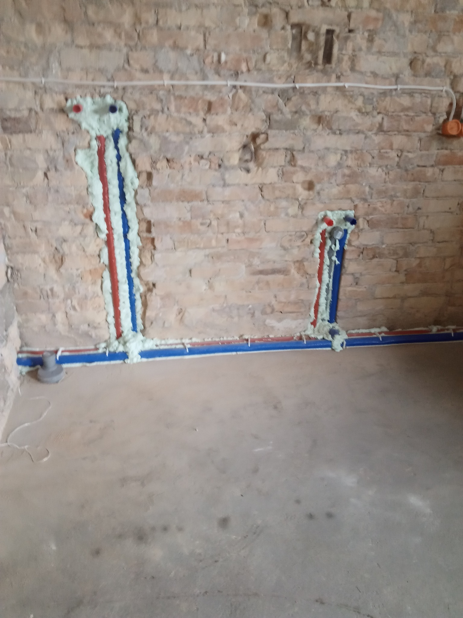 Miedziane i niebieskie rury wodne w ścianie z czerwonej cegły, zabezpieczone pianką montażową, widoczne na tle surowego betonu.