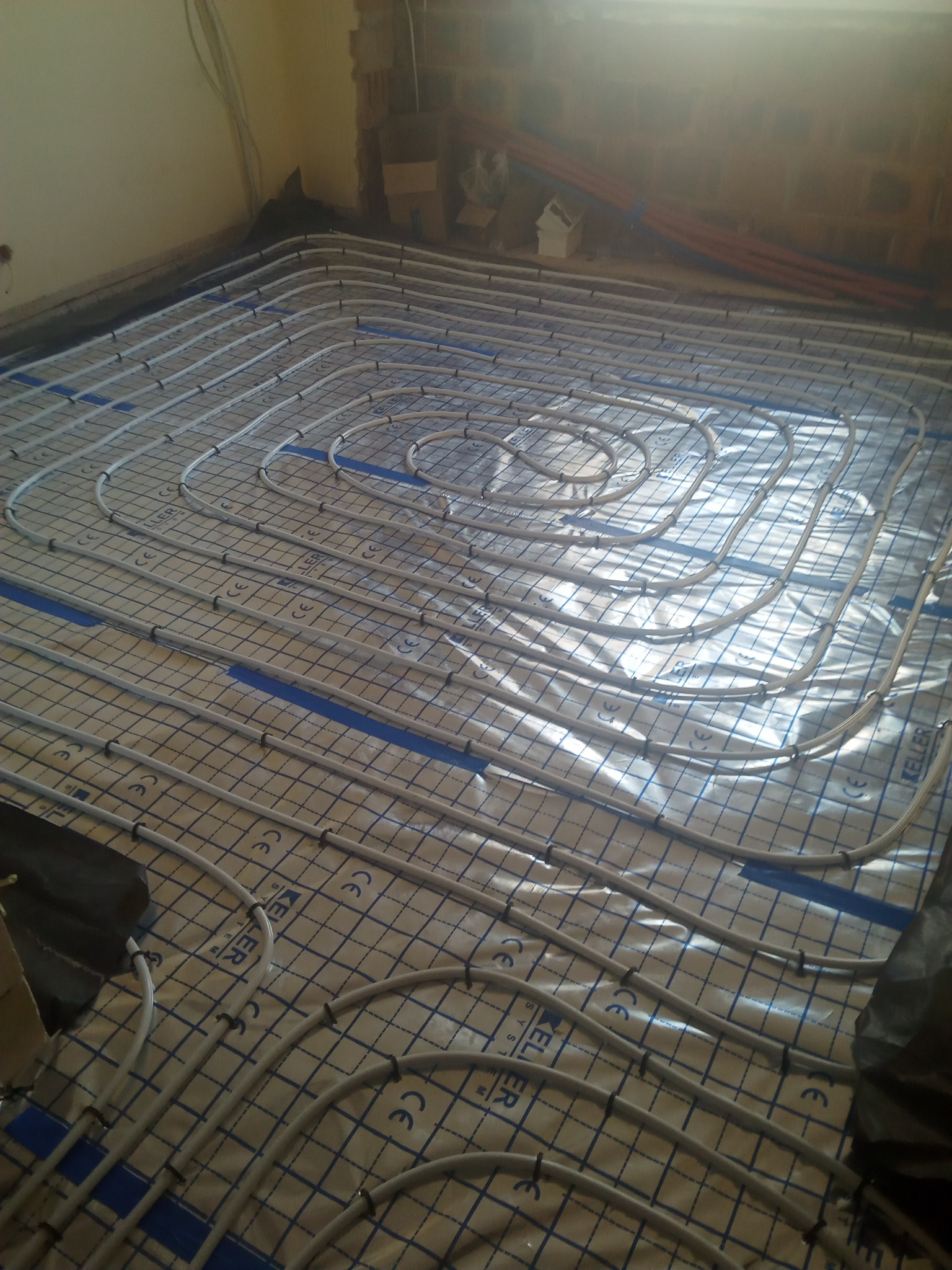 Rozłożone rury ogrzewania podłogowego na folii z nadrukiem kratki, zabezpieczone siatką i spinkami, widok z góry na etapie montażu instalacji w surowym stanie.